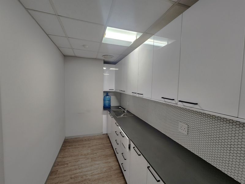 A clean, modern office kitchenette featuring white cabinetry, dark countertops, a stainless steel sink, and a stylish hexagonal tile backsplash.