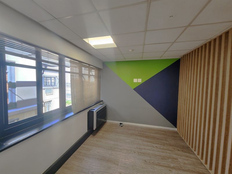 A bright, modern office space featuring wood-look flooring, slatted wood accent wall, suspended ceiling, and a wall-mounted air conditioning unit.