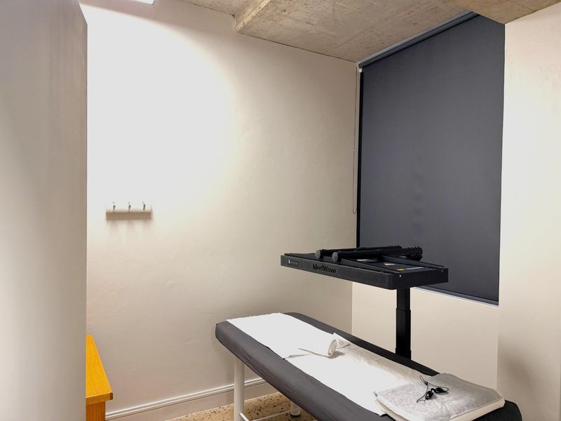 A functional office or treatment room featuring exposed concrete ceilings, terrazzo flooring, and a neutral wall finish with a fitted roller blind.