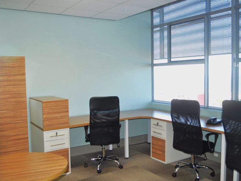 A functional office space featuring built-in desk workstations, carpeted flooring, and a large window with integrated blinds providing natural light.