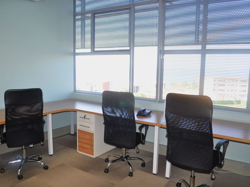 A functional office space featuring a large built-in desk with integrated storage, carpeted flooring, and expansive windows providing natural light.