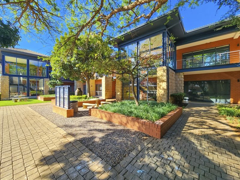 A well-maintained office complex courtyard featuring brick paving, stone-clad accents, and large glass facades surrounded by mature trees and landscaped garden beds.