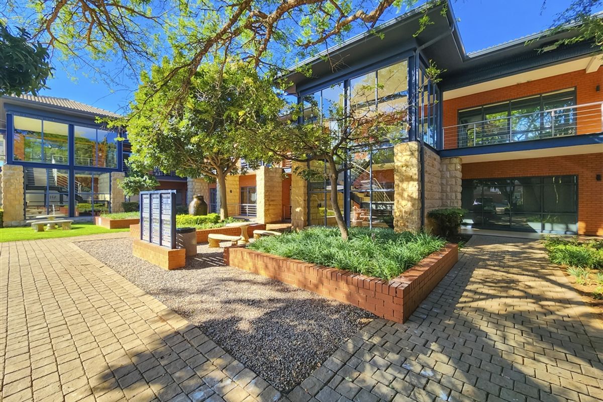A well-maintained office complex courtyard featuring brick paving, stone-clad accents, and large glass facades surrounded by mature trees and landscaped garden beds.