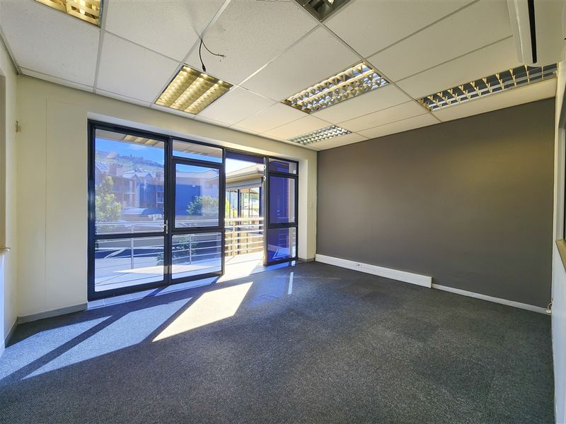 An empty office space featuring carpet flooring, a suspended ceiling with recessed lighting, and a glass door leading to an outdoor balcony area.