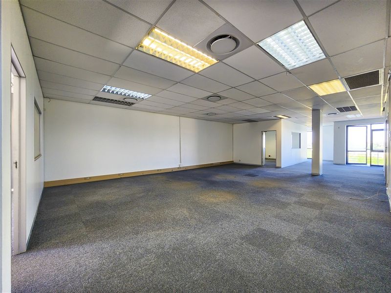 A spacious, open-plan office area featuring a suspended ceiling with recessed lighting, carpeted flooring, and large windows providing natural light.