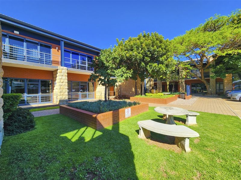 A well-maintained office courtyard featuring landscaped garden beds, paved walkways, and modern brick and glass building facades with balcony access.