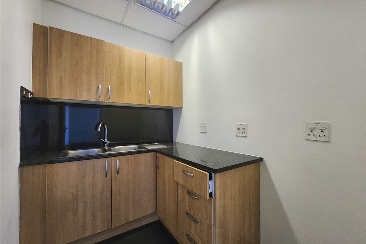 A compact office kitchenette featuring wood-finish cabinetry, granite countertops, a stainless steel sink, and a modern black glass splashback.