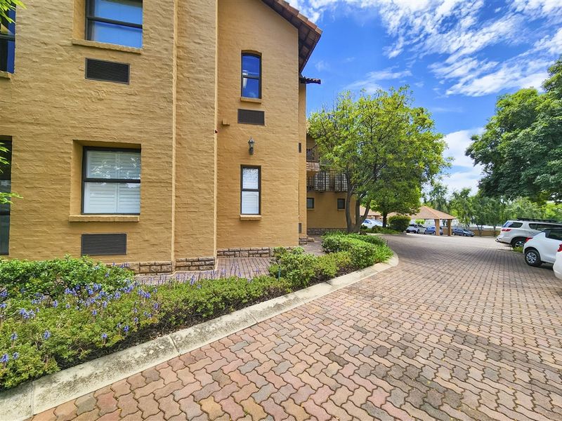 Exterior view of the office building featuring a textured stucco facade, terracotta roof tiles, and a well-maintained brick paved driveway with landscaped garden beds.