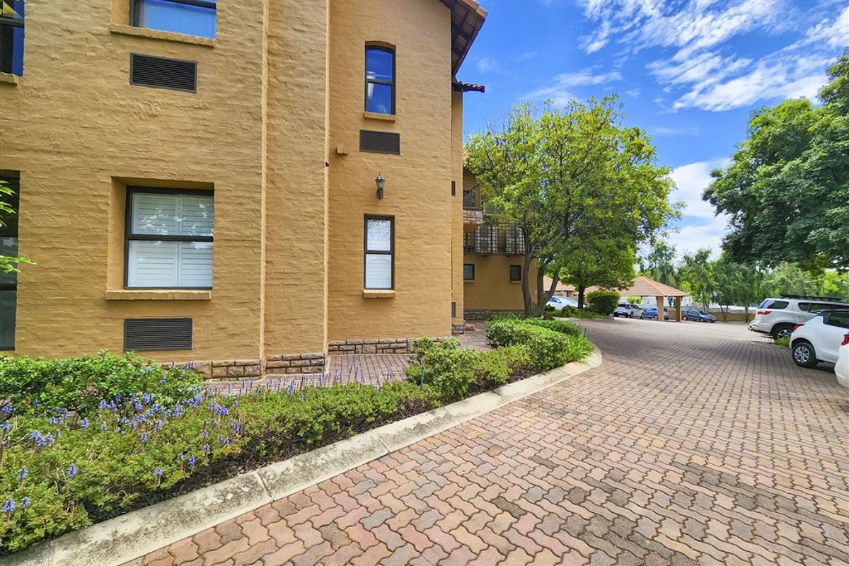 Exterior view of the office building featuring a textured stucco facade, terracotta roof tiles, and a well-maintained brick paved driveway with landscaped garden beds.