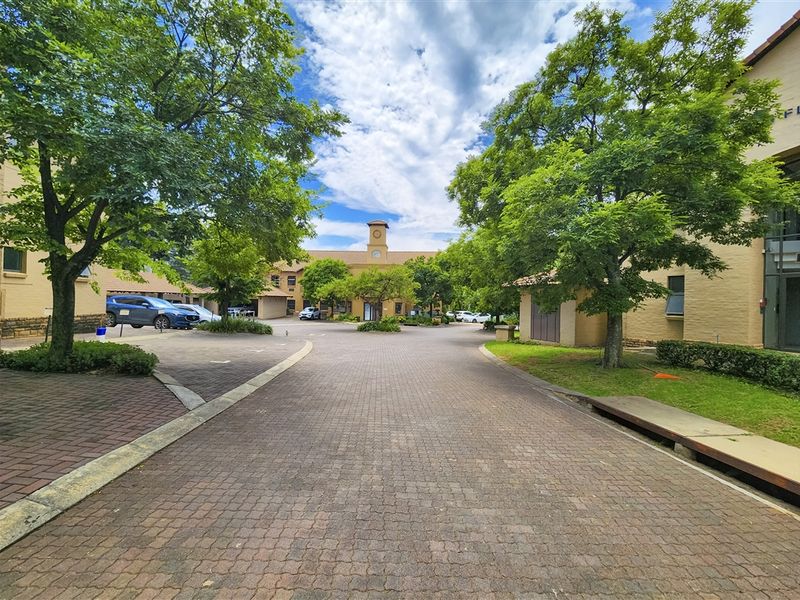 A well-maintained office park entrance featuring a paved driveway, mature trees, and landscaped gardens leading to the main building facade.