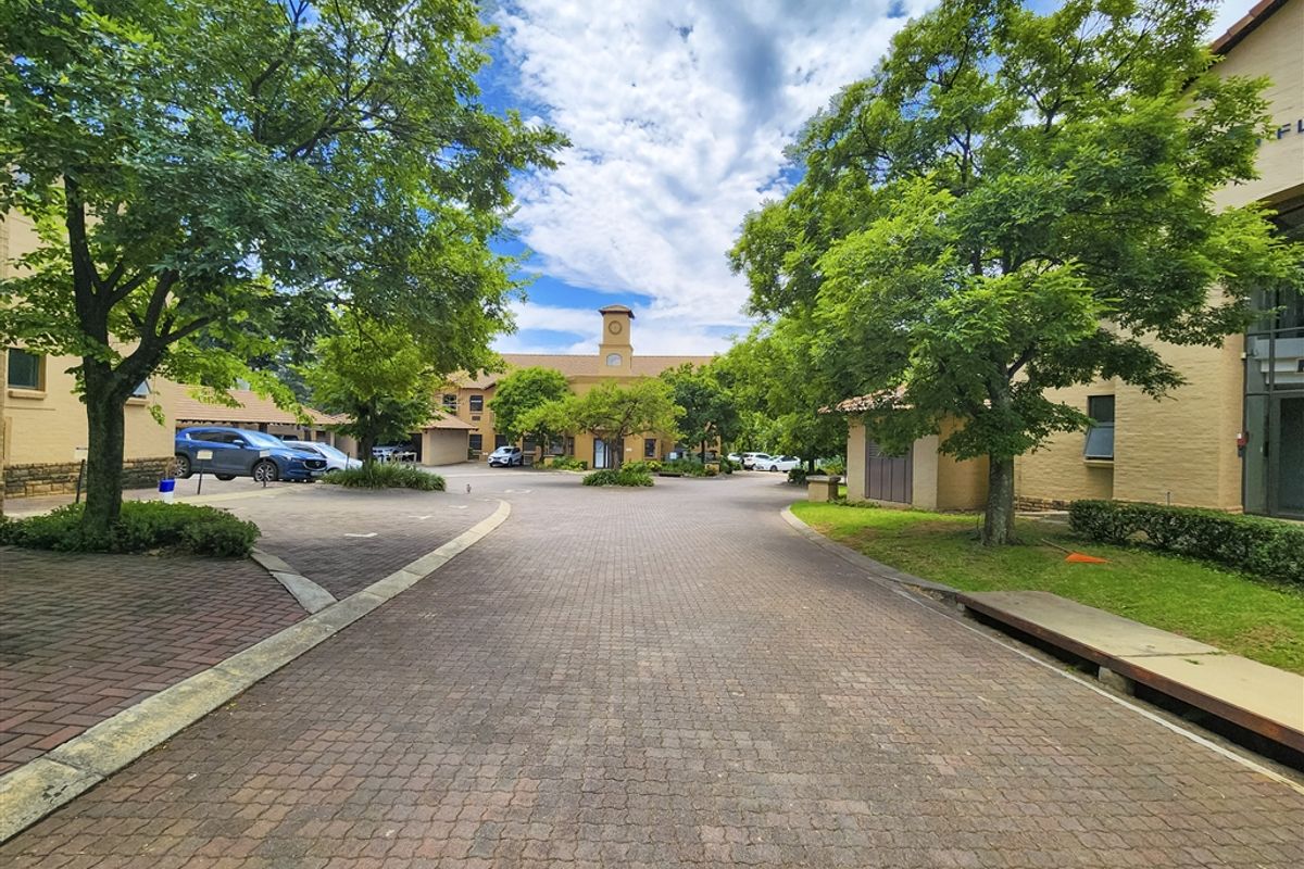 A well-maintained office park entrance featuring a paved driveway, mature trees, and landscaped gardens leading to the main building facade.
