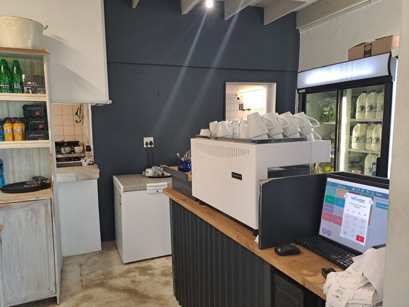 A functional commercial kitchen area featuring a coffee station, display refrigeration, built-in shelving, and exposed ceiling beams.