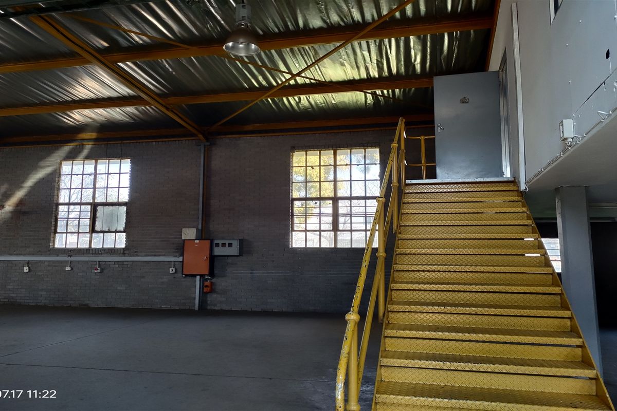 Spacious industrial warehouse area featuring a prominent yellow metal staircase, exposed steel roof trusses, concrete flooring, and large industrial windows providing natural light.