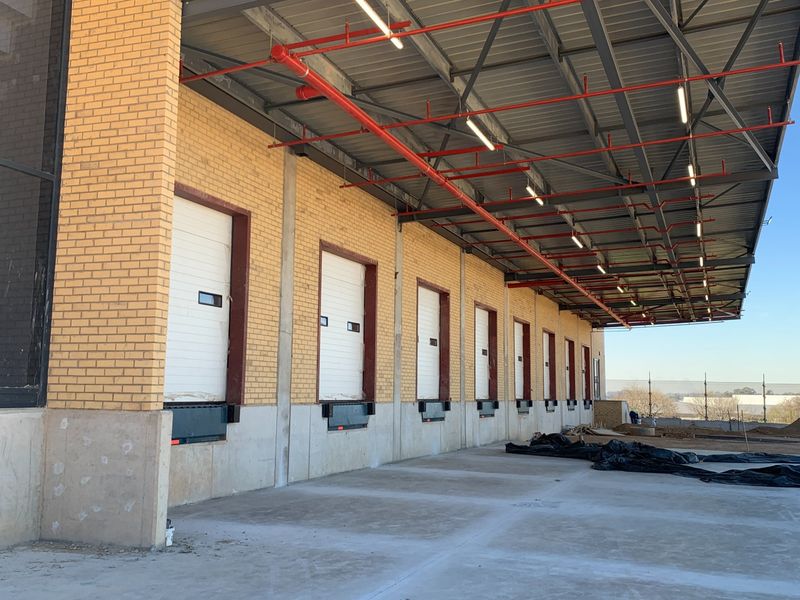 Industrial loading bay area featuring multiple dock doors, dock levelers, and an expansive covered concrete apron with exposed steel roof structure and fire sprinklers.