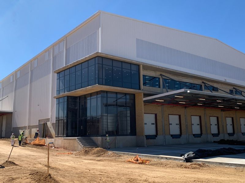 Modern industrial warehouse facility featuring extensive metal cladding, a prominent glass-fronted office section, and multiple loading docks with roller shutter doors.