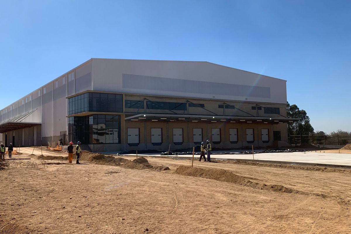 A modern industrial warehouse facility featuring multiple loading docks, extensive metal cladding, and large glass facade elements under construction.