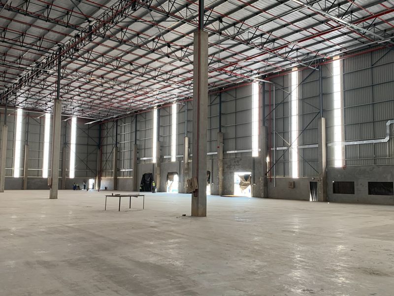 Expansive industrial warehouse space featuring high ceilings with exposed steel trusses, fire sprinkler systems, concrete flooring, and natural light through vertical wall panels.