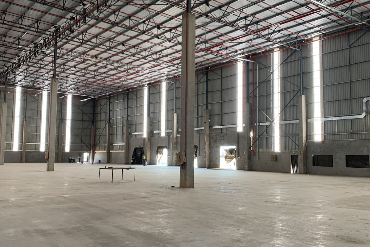 Expansive industrial warehouse space featuring high ceilings with exposed steel trusses, fire sprinkler systems, concrete flooring, and natural light through vertical wall panels.