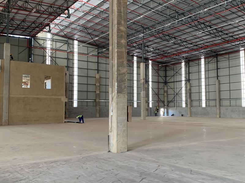 Large industrial warehouse space featuring high ceilings with exposed steel trusses, concrete columns, and fire sprinkler systems, currently under construction.