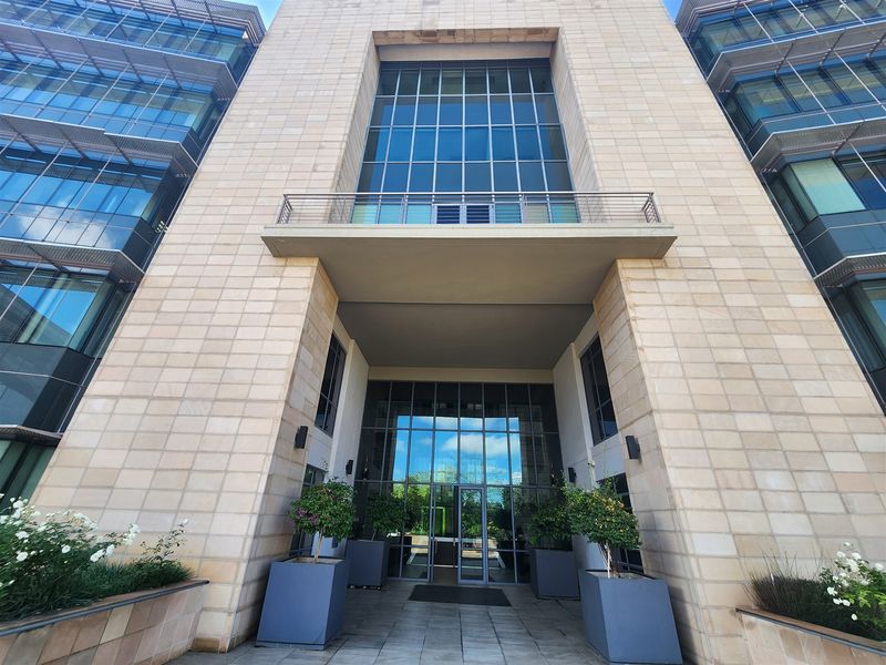 Modern office building entrance featuring stone facade, large glass curtain walls, and a prominent balcony above the main glass entry doors.