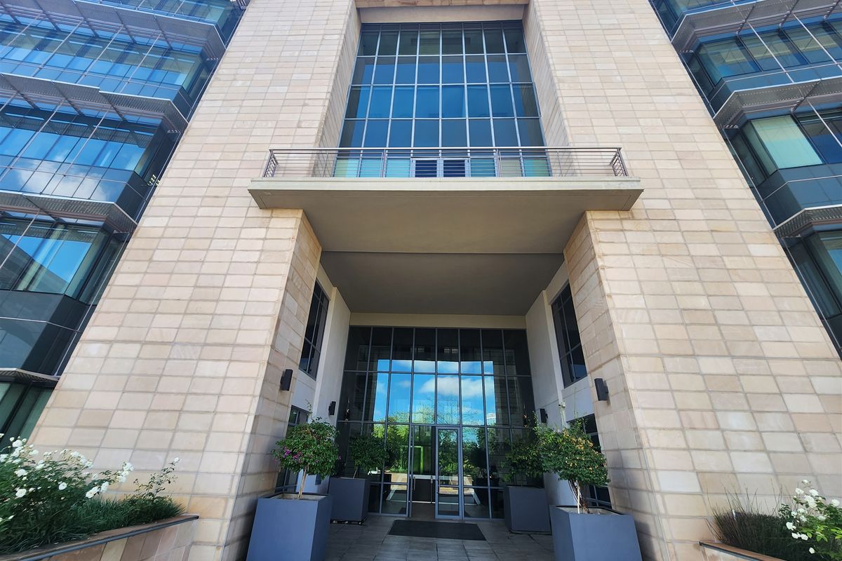 Modern office building entrance featuring stone facade, large glass curtain walls, and a prominent balcony above the main glass entry doors.