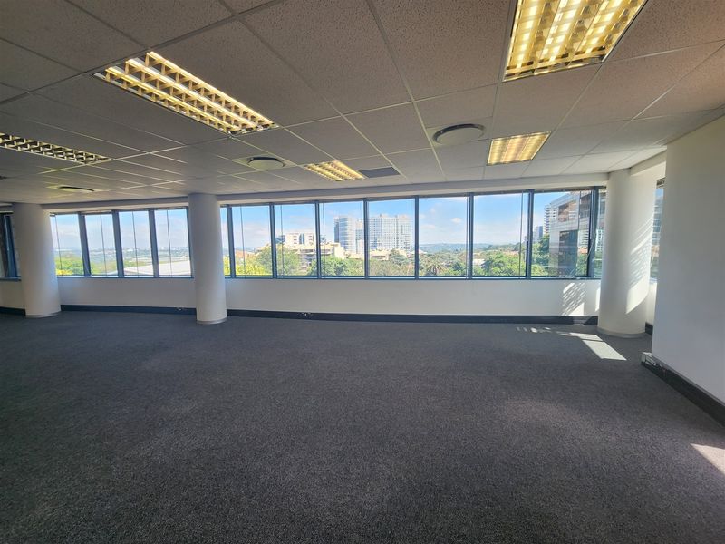 A spacious, empty open-plan office area featuring large windows with city views, carpeted flooring, and a suspended ceiling with recessed lighting.