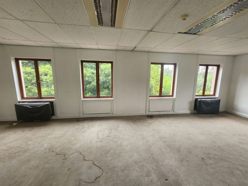 An empty office space featuring large timber-framed windows, suspended ceilings with recessed lighting, and HVAC units, requiring flooring and ceiling refurbishment.