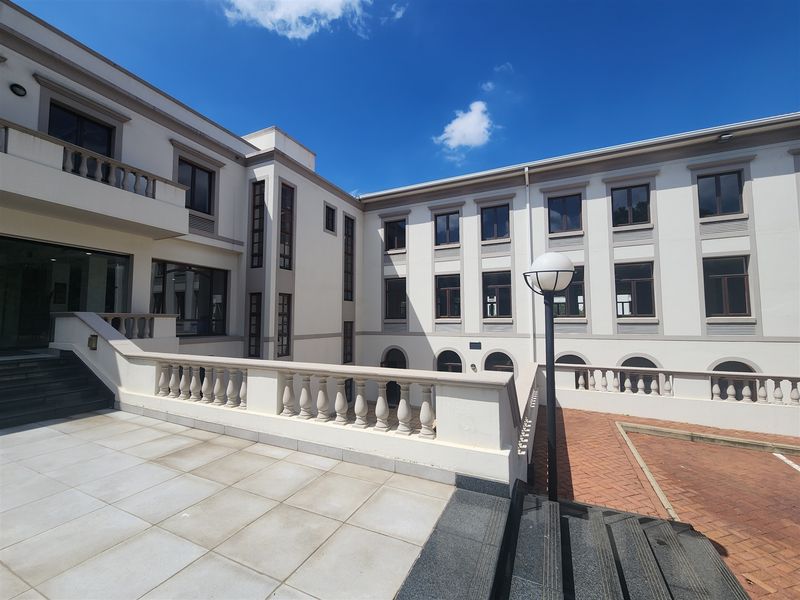 A well-maintained multi-story office building exterior featuring a paved terrace, balustrade detailing, and an external staircase leading to the courtyard area.
