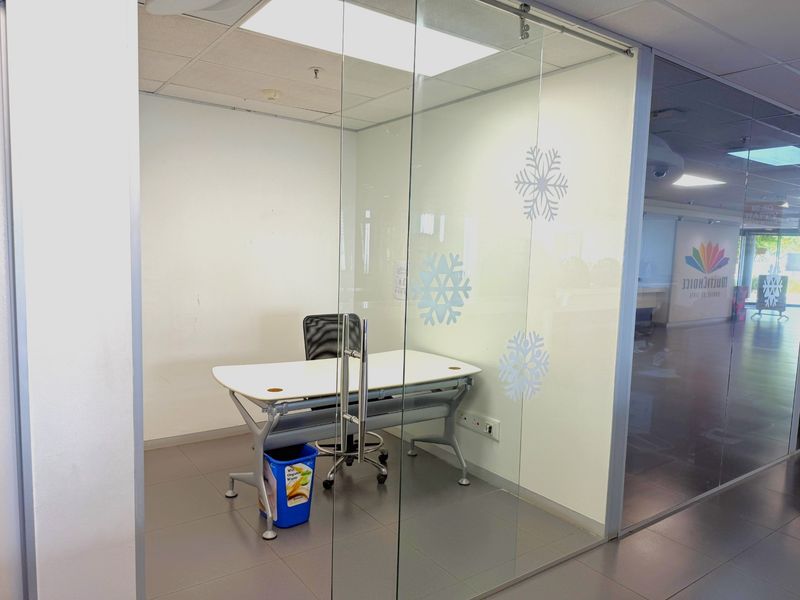 A compact, modern office space featuring a glass partition wall, tiled flooring, and a suspended ceiling with recessed lighting.