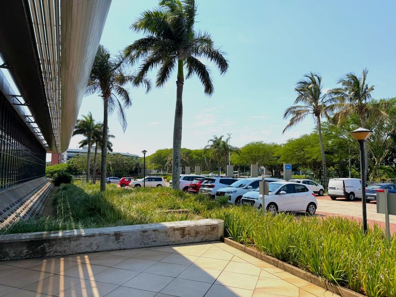 Exterior view of the office building facade featuring a paved walkway, landscaped garden area, and adjacent parking facilities.