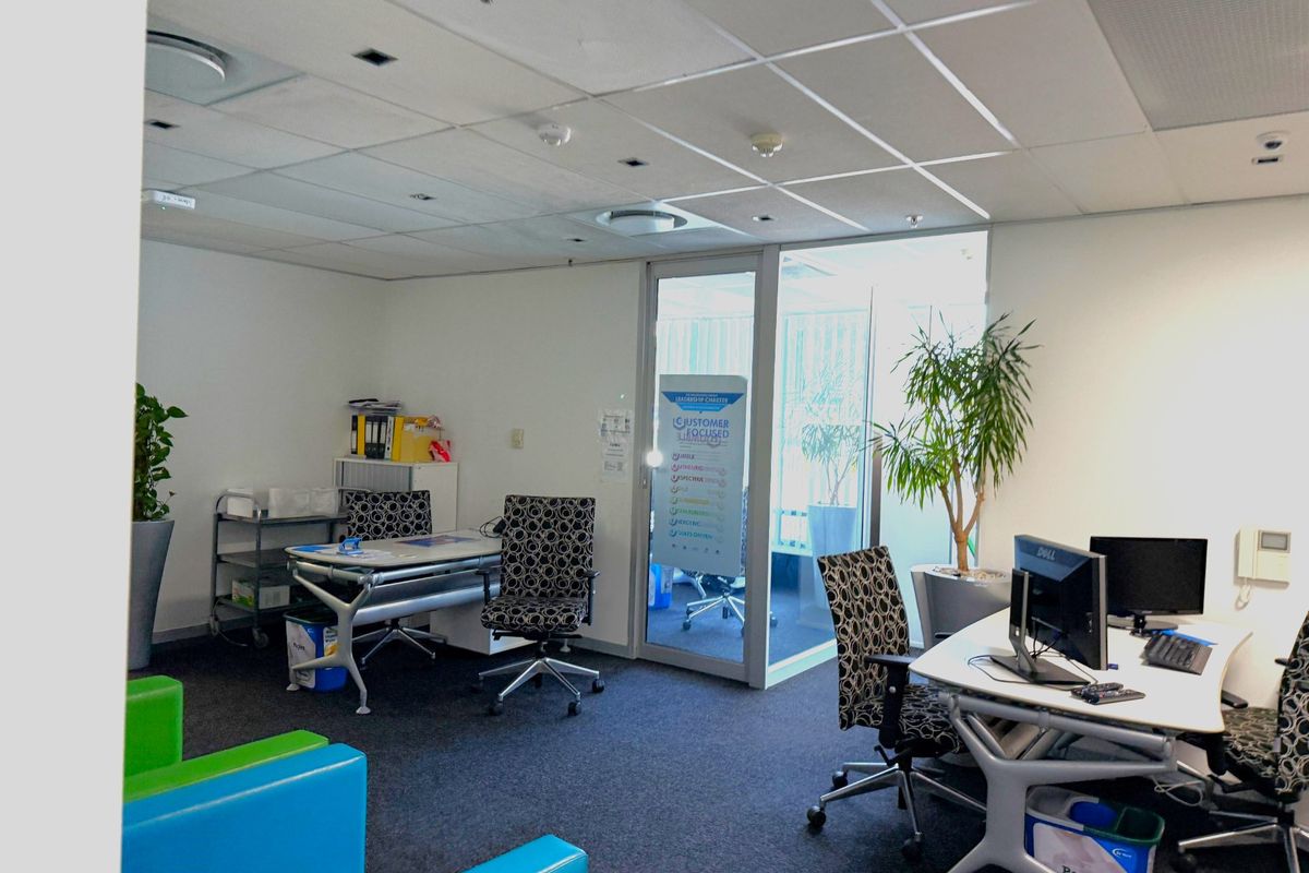 A functional office space featuring a suspended ceiling, recessed lighting, carpeted flooring, and a glass partition door leading to an adjacent area.