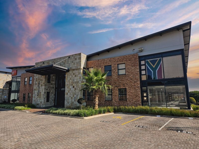 Modern office building exterior featuring a combination of stone and face brick facade, large glass windows, and a paved parking area.