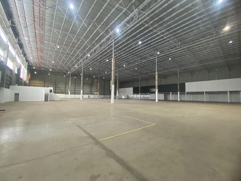 A large, open-plan industrial warehouse space featuring high ceilings with exposed steel trusses, concrete flooring, and high-bay lighting, suitable for logistics or manufacturing.