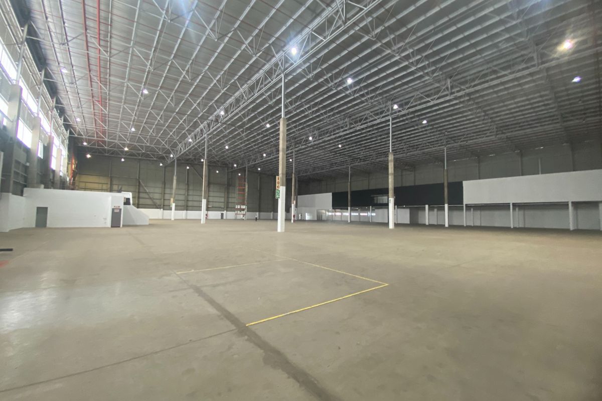 A large, open-plan industrial warehouse space featuring high ceilings with exposed steel trusses, concrete flooring, and high-bay lighting, suitable for logistics or manufacturing.