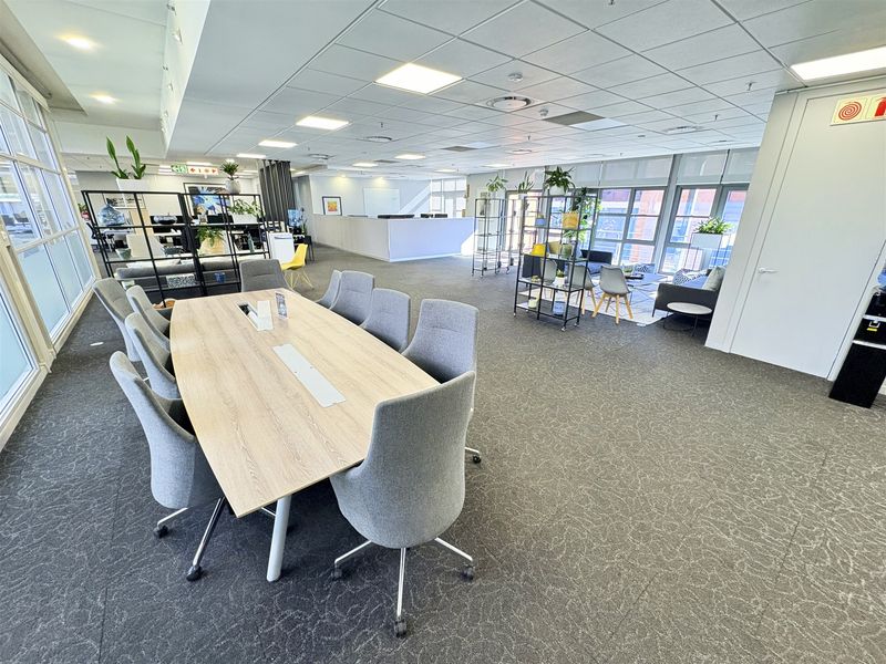 Spacious open-plan office area featuring a large meeting table, reception desk, modern suspended ceiling, and ample natural light from large windows.