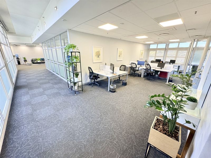 A bright, open-plan office space featuring modern glass partitions, suspended ceilings with recessed lighting, and large windows providing ample natural light.
