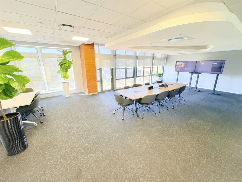 A modern, well-lit office boardroom featuring large windows, carpeted flooring, an exposed brick accent column, and a suspended ceiling with integrated climate control.