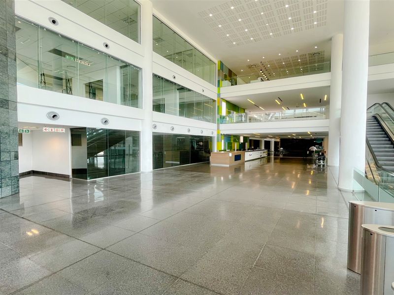 Spacious modern office building atrium featuring polished stone floors, glass-walled office suites, a central reception area, and an escalator for multi-level access.