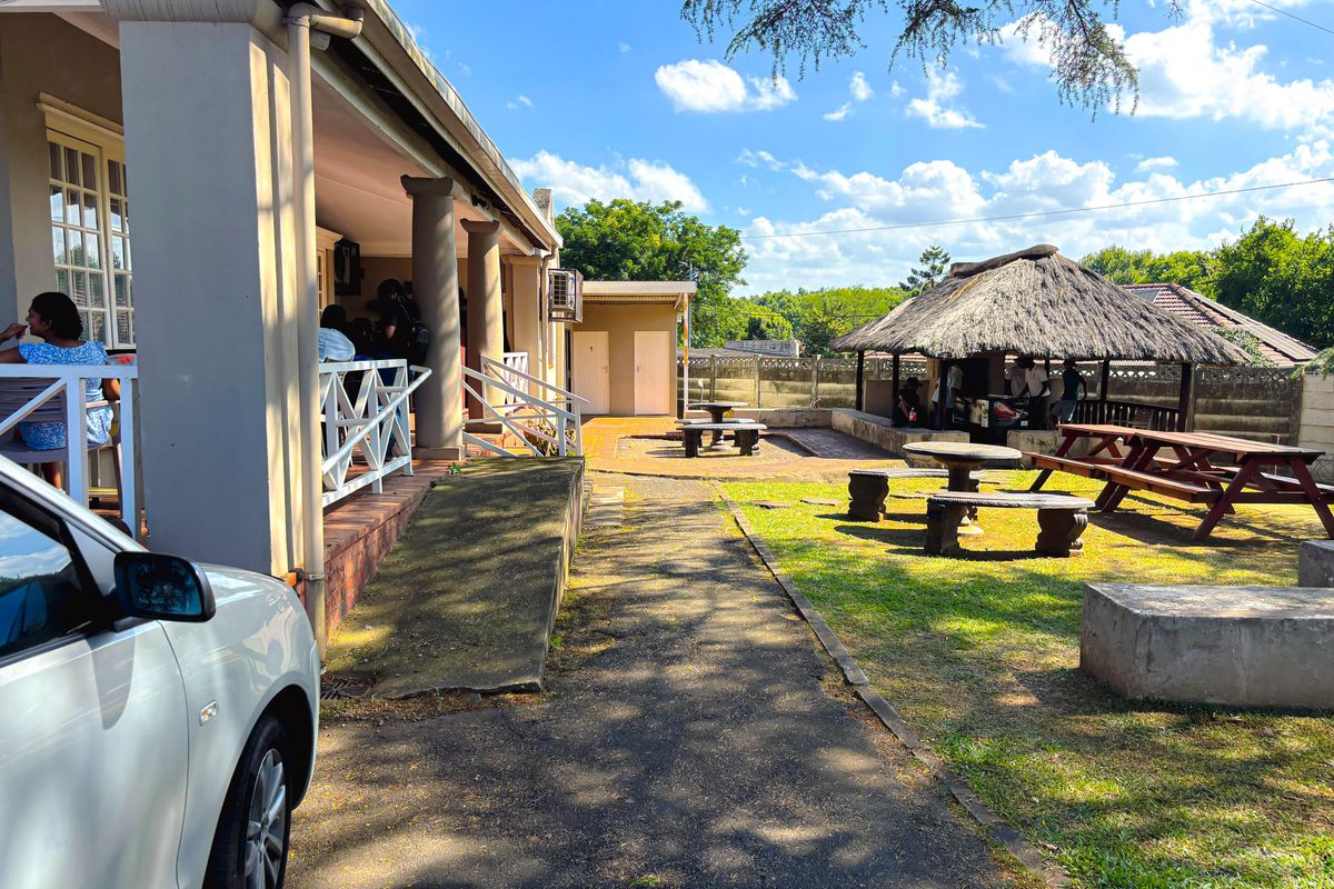 Rear exterior view of the commercial property featuring a covered patio, accessible concrete ramp, and a detached thatched lapa area set within a garden space.