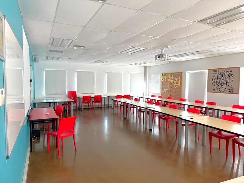 A spacious office or training room featuring a suspended ceiling, recessed lighting, ceiling-mounted projector, and large windows with roller blinds.