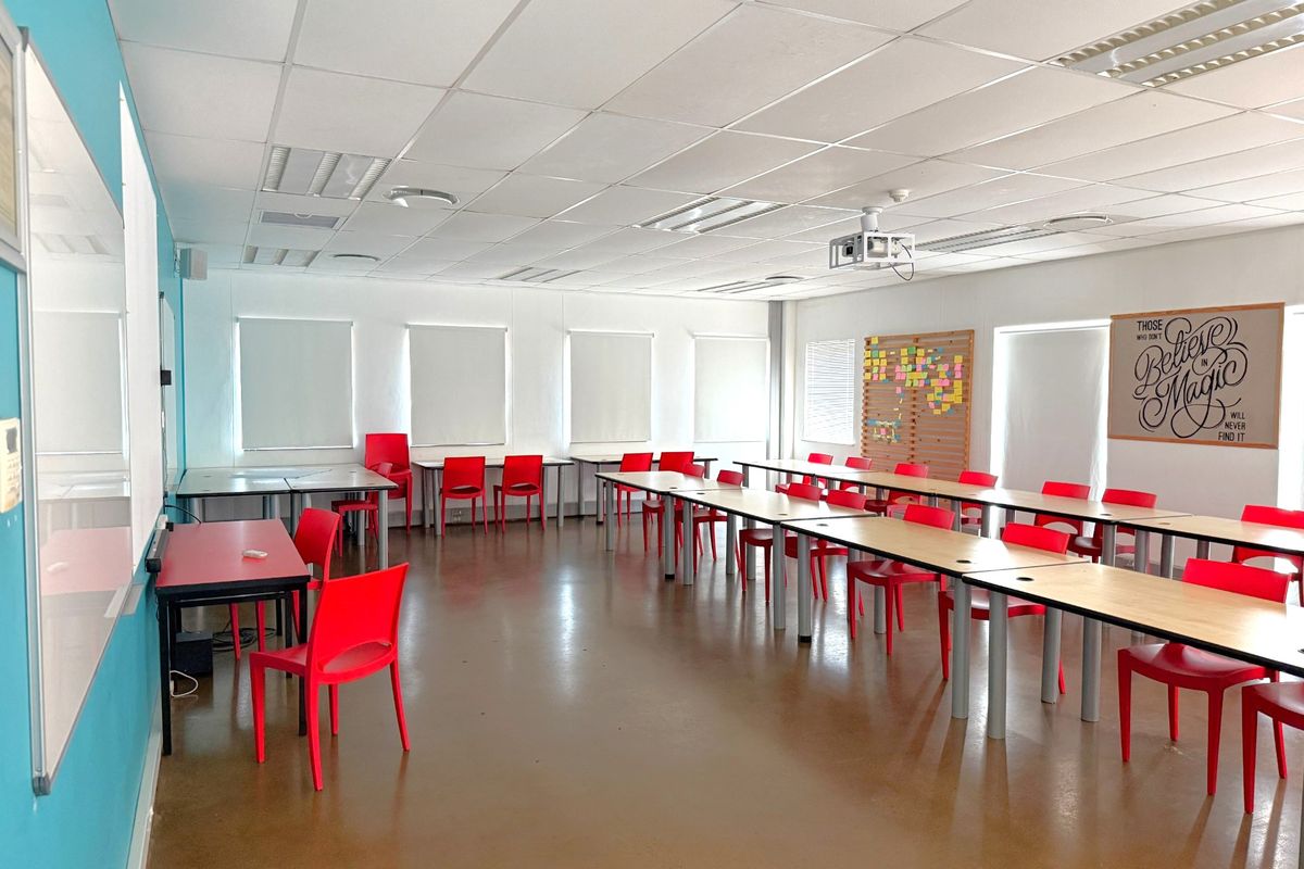 A spacious office or training room featuring a suspended ceiling, recessed lighting, ceiling-mounted projector, and large windows with roller blinds.