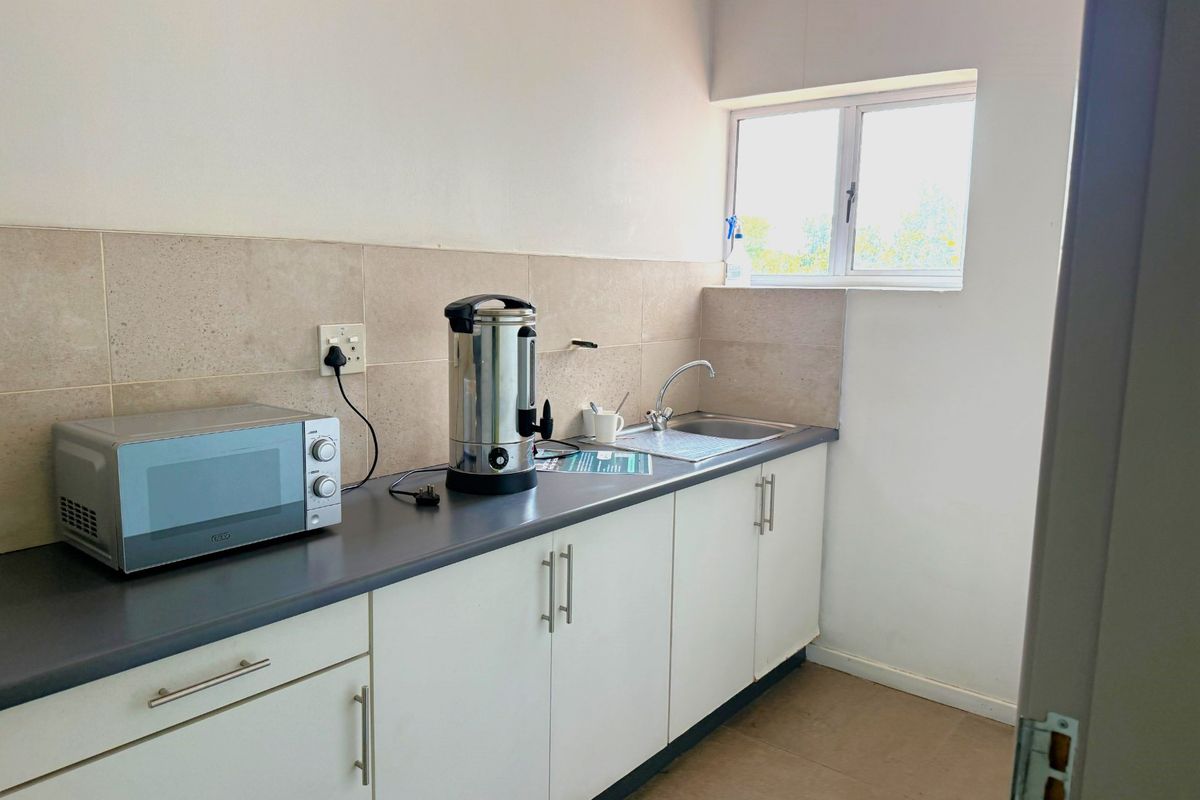 A functional office kitchenette featuring white built-in cabinetry, a laminate countertop, a stainless steel sink, and a neutral tiled backsplash.