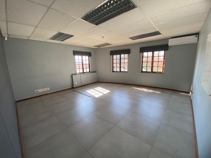 A bright, empty office space featuring tiled flooring, a suspended ceiling with recessed lighting, and a wall-mounted air conditioning unit.