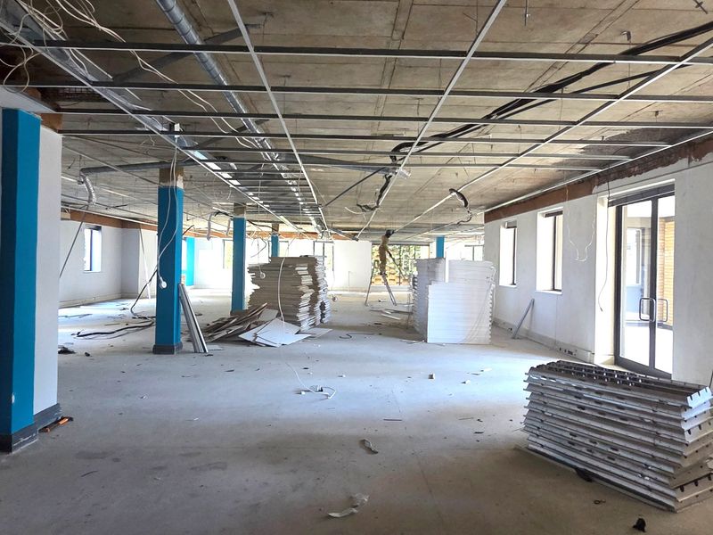 A large, open-plan office space currently under renovation, featuring an exposed ceiling grid, structural columns, and large windows providing ample natural light.