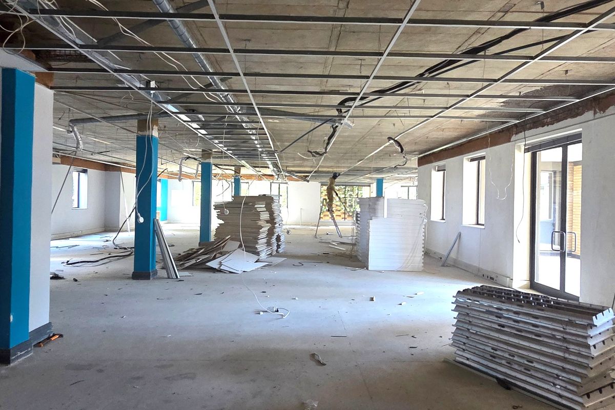 A large, open-plan office space currently under renovation, featuring an exposed ceiling grid, structural columns, and large windows providing ample natural light.