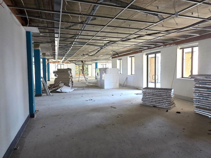 An open-plan office space currently under renovation, featuring exposed structural elements, a suspended ceiling grid, and large windows providing natural light.