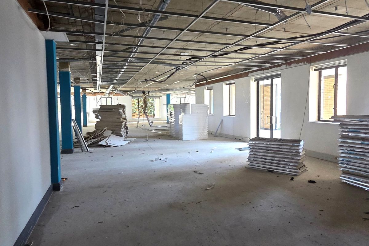 An open-plan office space currently under renovation, featuring exposed structural elements, a suspended ceiling grid, and large windows providing natural light.