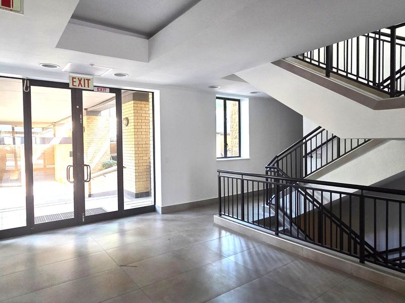 A bright and modern office building entrance lobby featuring tiled flooring, a prominent staircase with metal balustrades, and large glass doors.