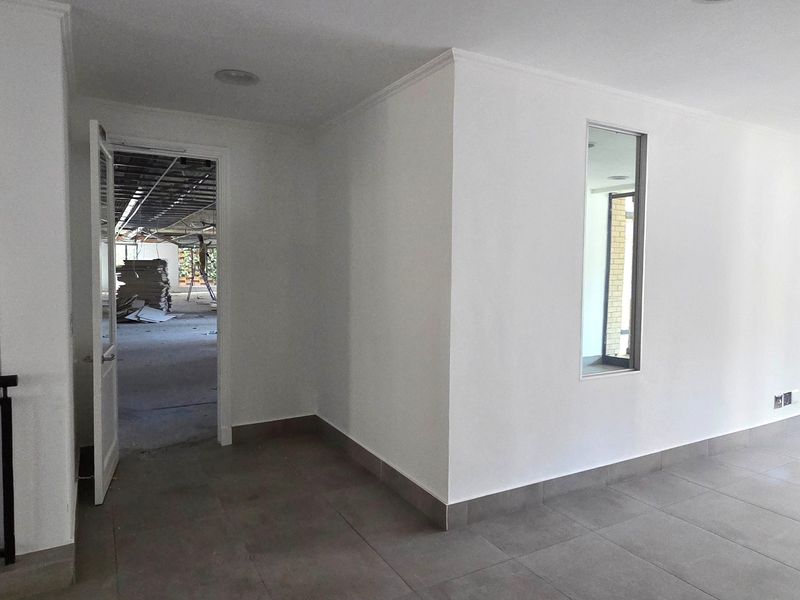 A bright, empty office space featuring neutral tiled flooring, recessed lighting, and an internal window looking into an adjacent area under renovation.