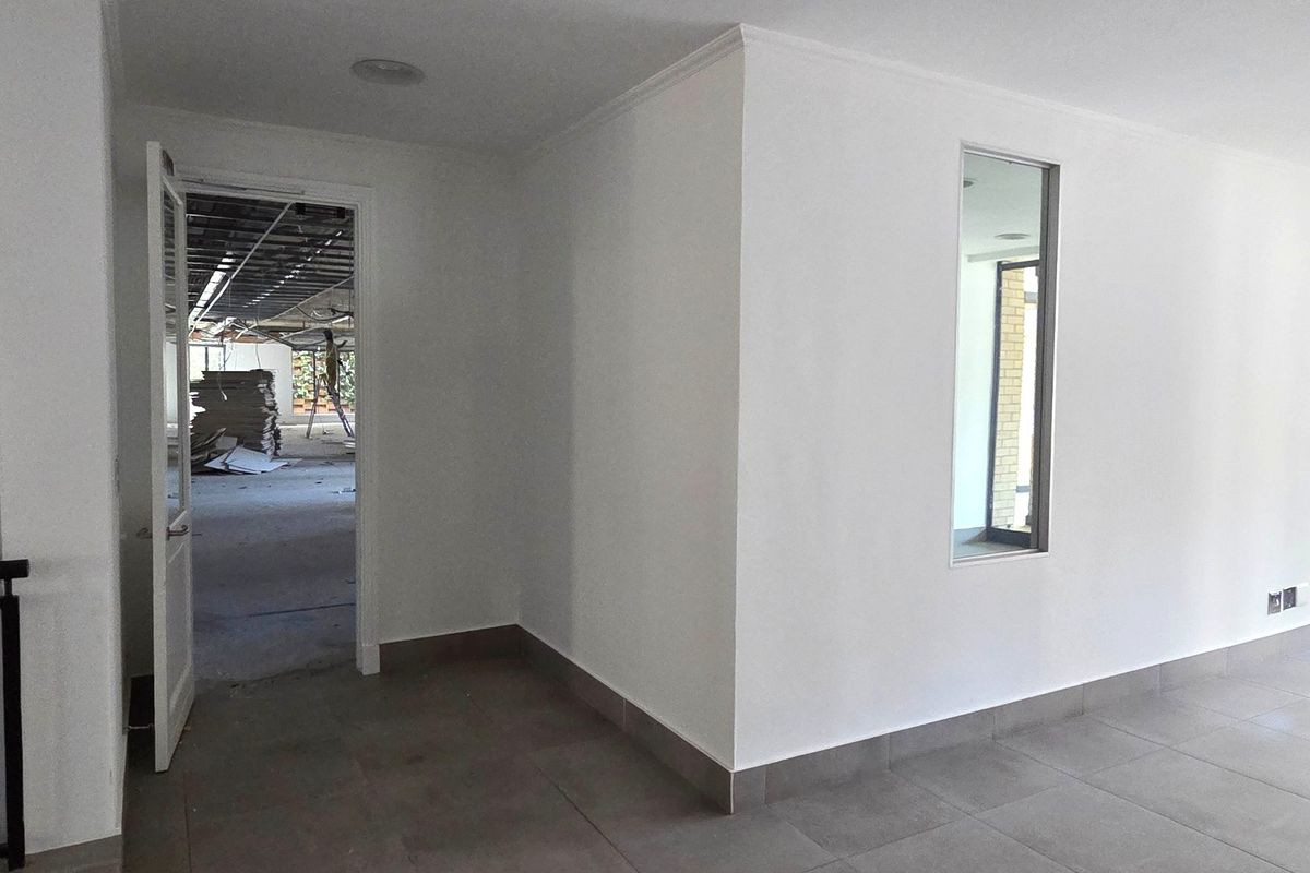 A bright, empty office space featuring neutral tiled flooring, recessed lighting, and an internal window looking into an adjacent area under renovation.